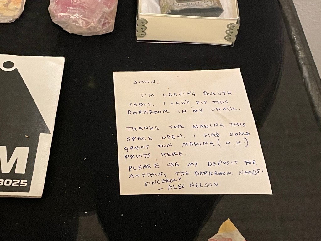 A close-up of a letter in the display. It says "John, I'm leaving Duluth. Sadly, I can't fit this darkroom in my Uhaul. Thanks for making this space open. I had some great fun malking (o.k.) prints here. Please use my deposit for anything the darkroom needs! Sincerely - Alex Nelson."